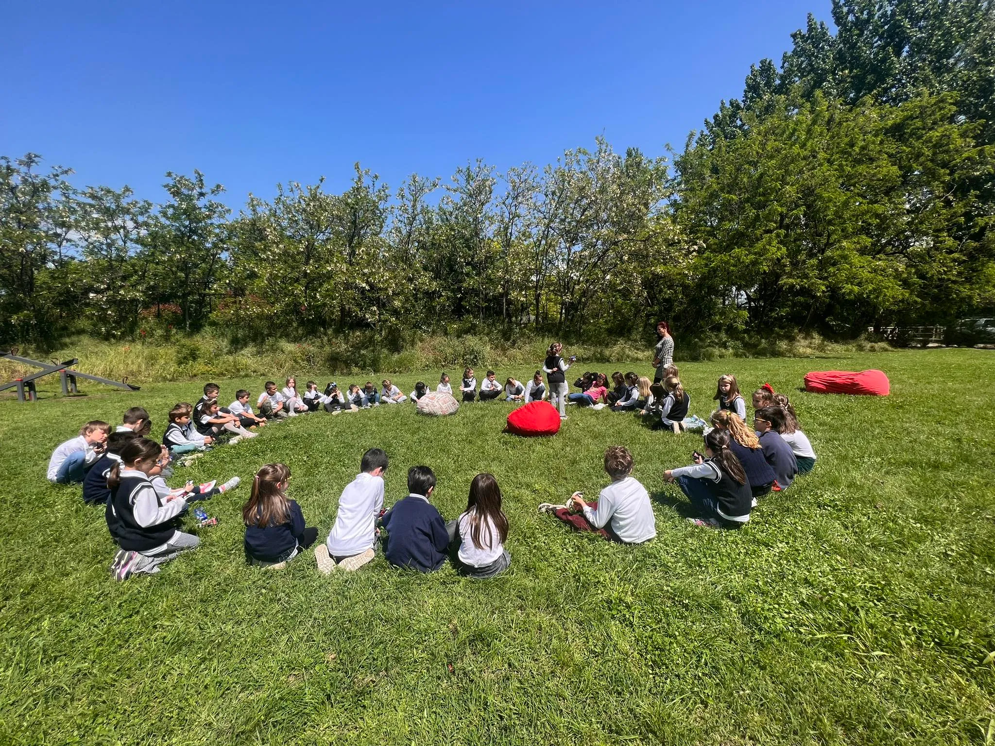 A large circle of uniformed children sitting on the grass with their teacher