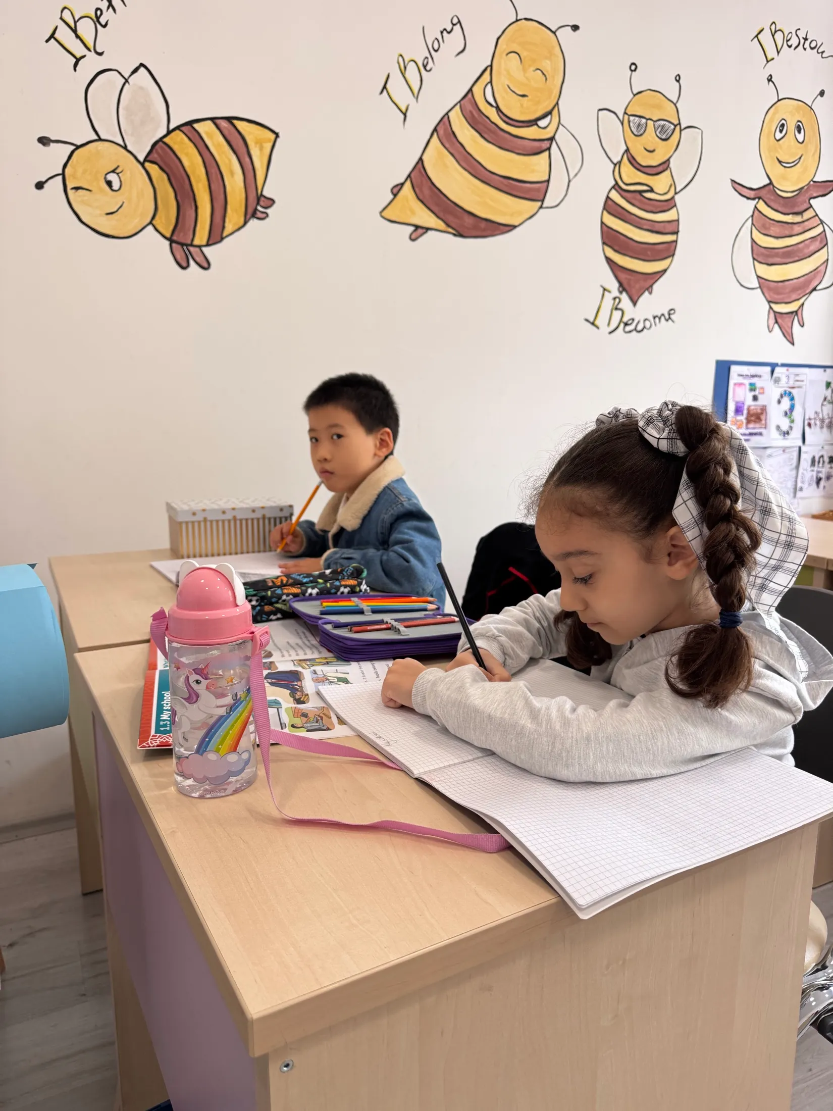 Two first graders writing at their desks beneath a hand-painted mural of bees labelled ‘I Belong’, ‘I Become’, and ‘I Bestow’