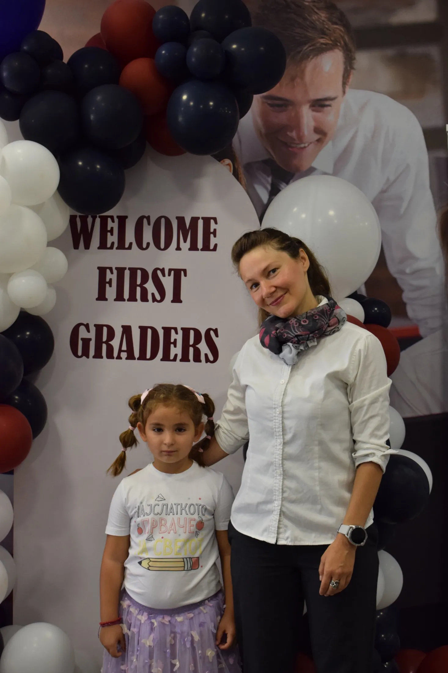 A first grader and her teacher in front of a “Welcome First Graders” balloon arch
