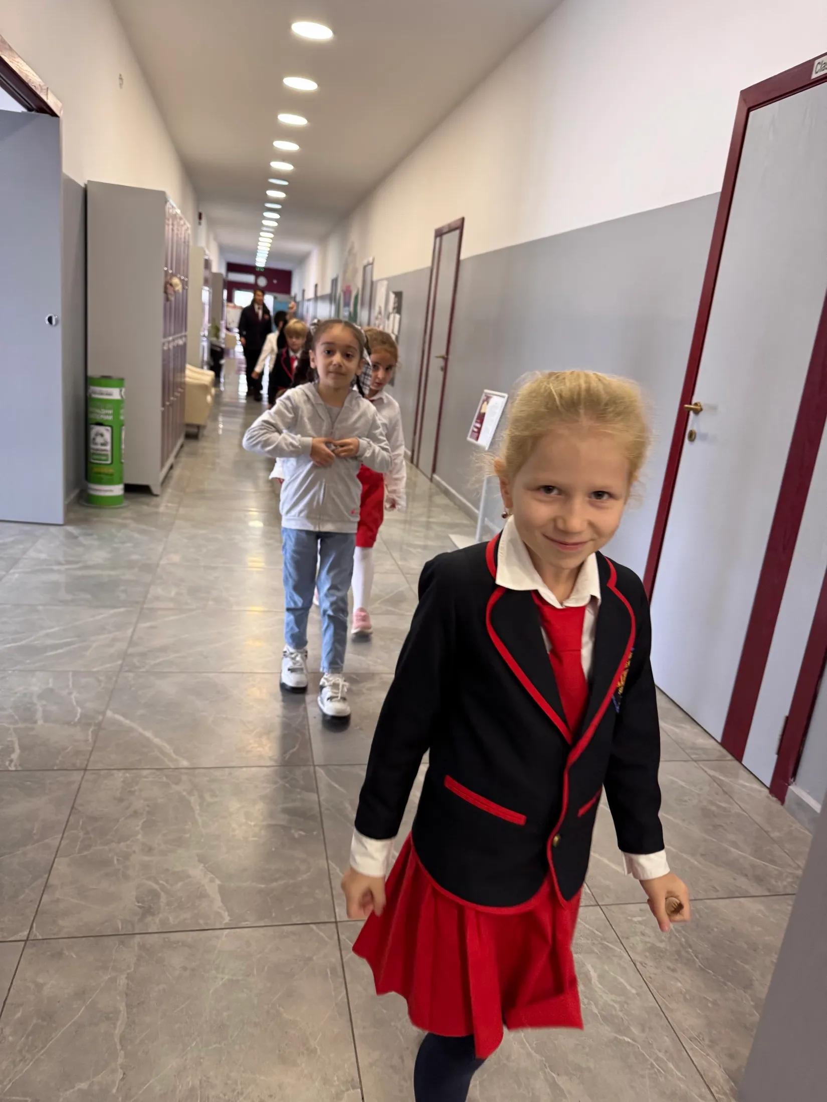 A smiling first grader in uniform leading her classmates down the school hallway